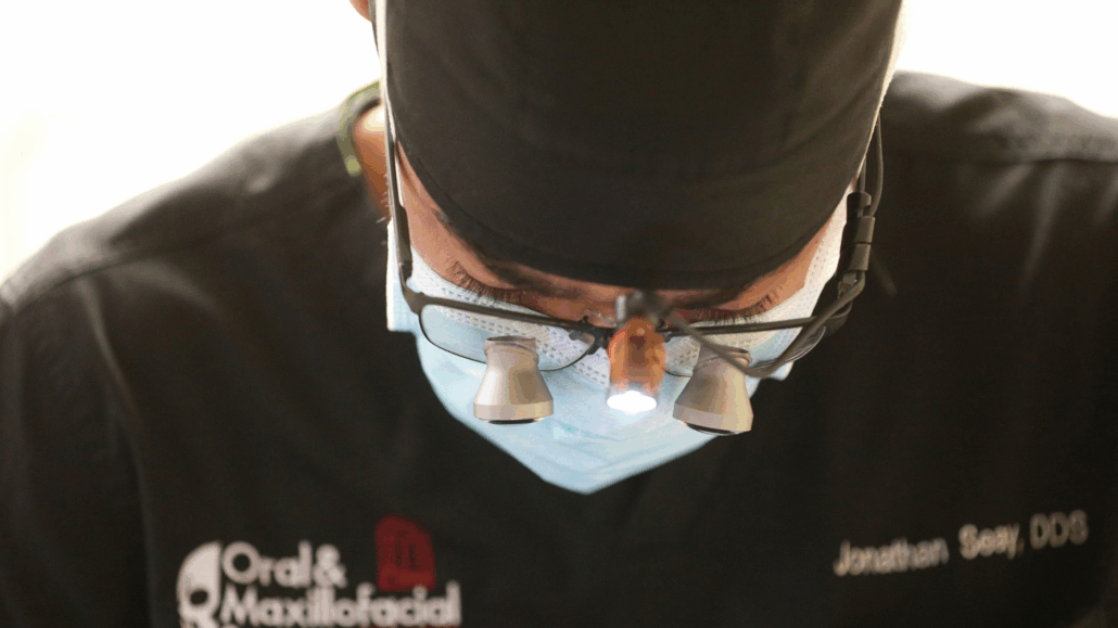 A person in a black mask and glasses, possibly a dentist, looking down at a microscope.