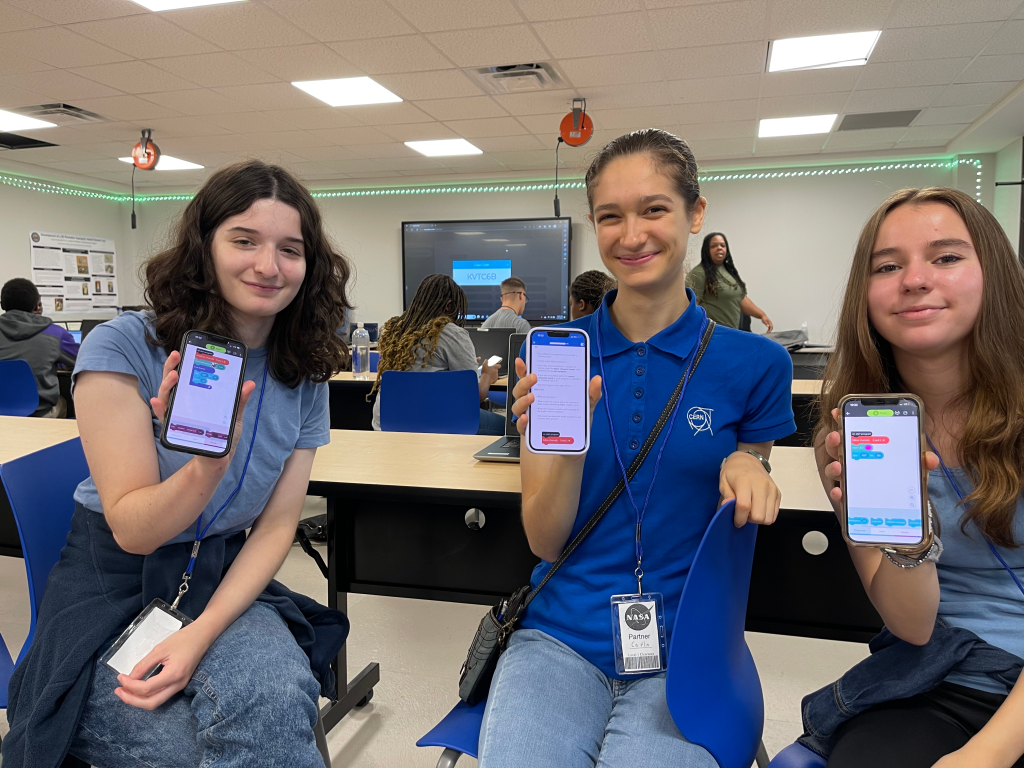 Three people holding up smartphones in a classroom setting.