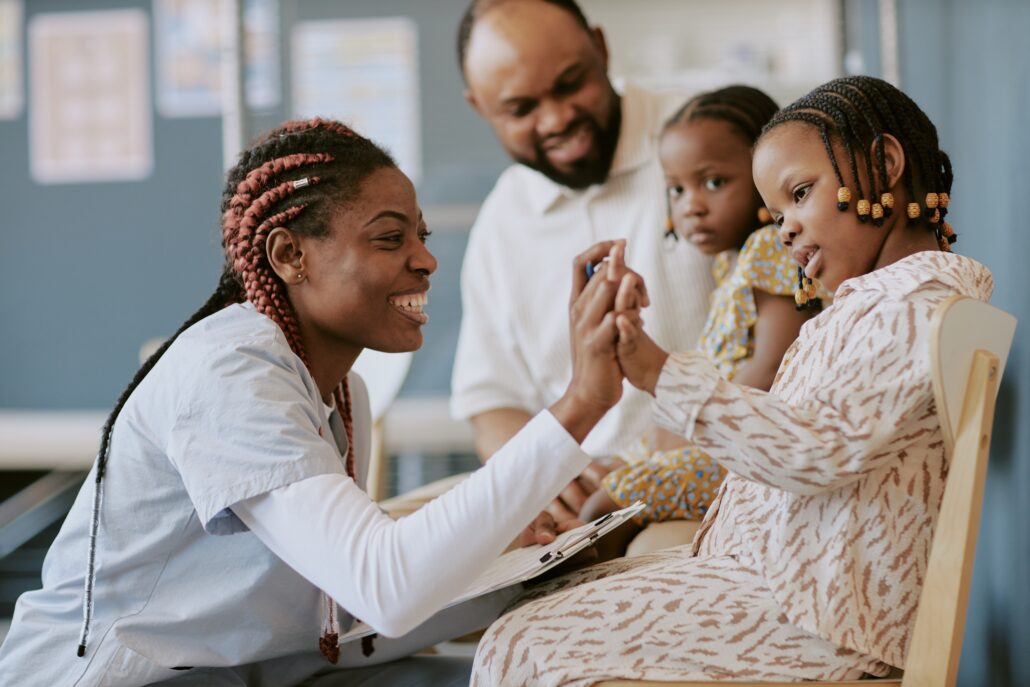 A woman in a white shirt and black pants is holding a child's hand, smiling at the camera. They are in a room with a group of people.