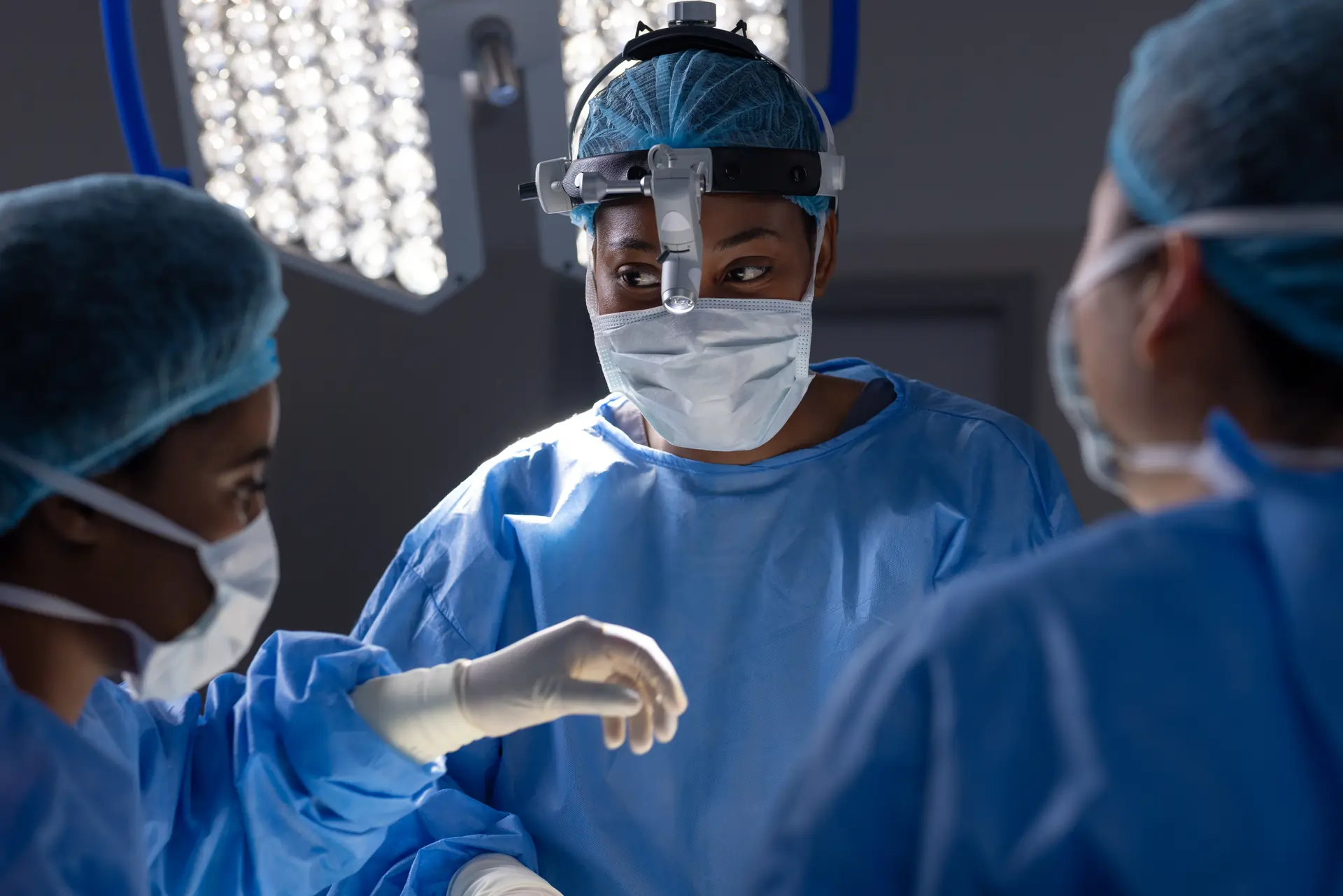 Medical team in operating room. Surgeon in blue gown and mask. Three nurses in blue scrubs. Surgeon in white gown and mask.