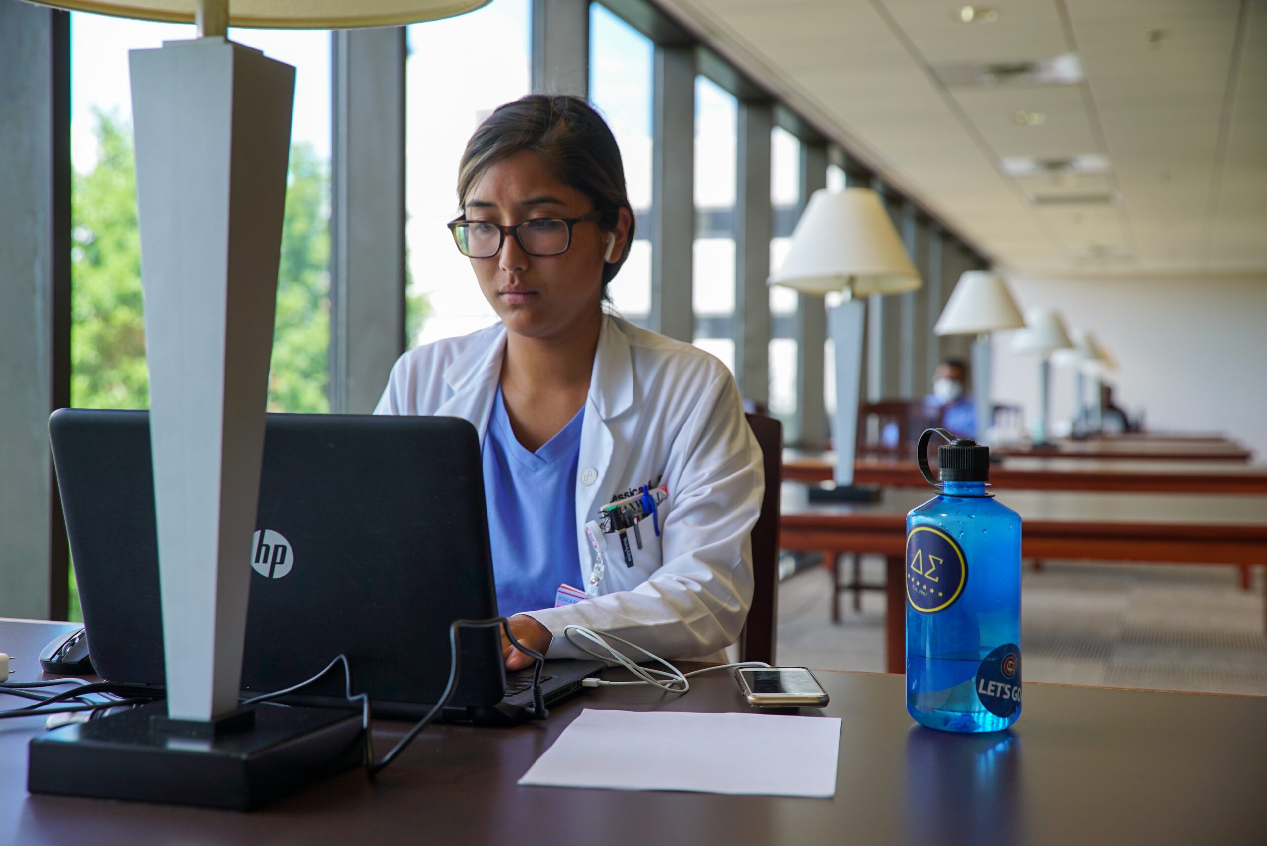 Woman in white coat sitting at table with laptop, working in library.