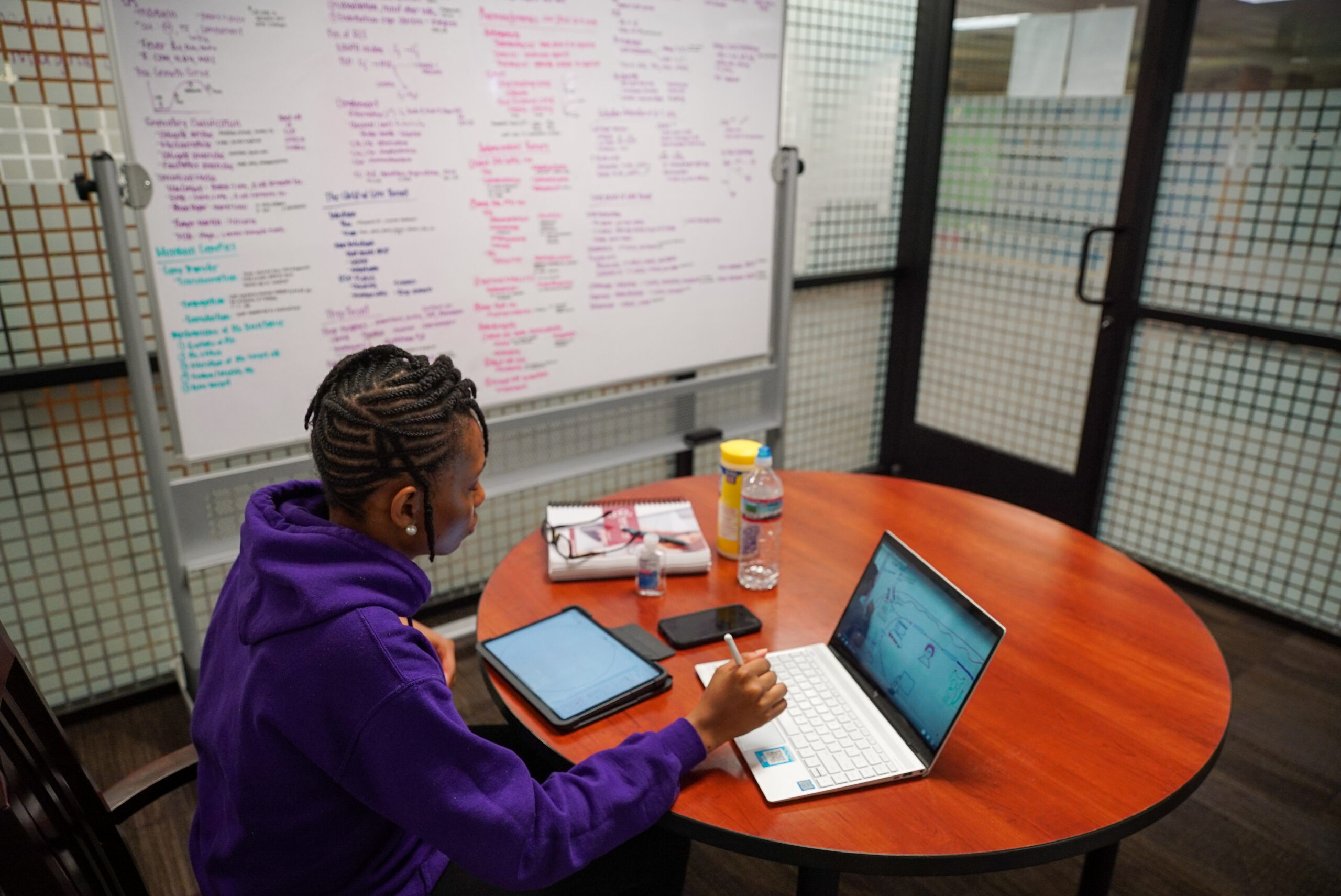 Person sitting at table with laptop, surrounded by whiteboard with writing.