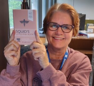 A woman holding a book titled "Squid's Little Pink Book" in front of a desk.