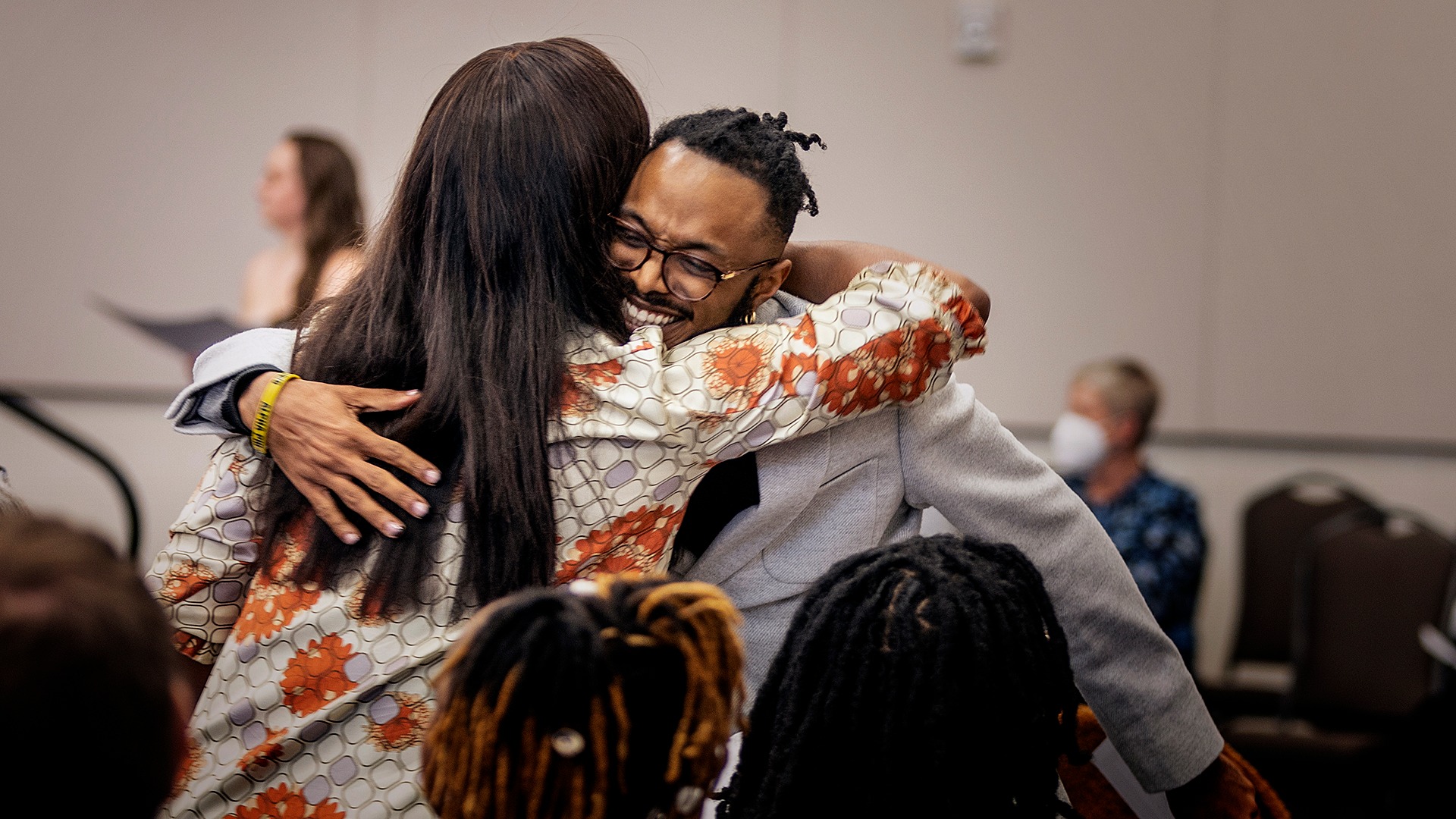 Two people hugging, one woman and one man, in a room with other people.