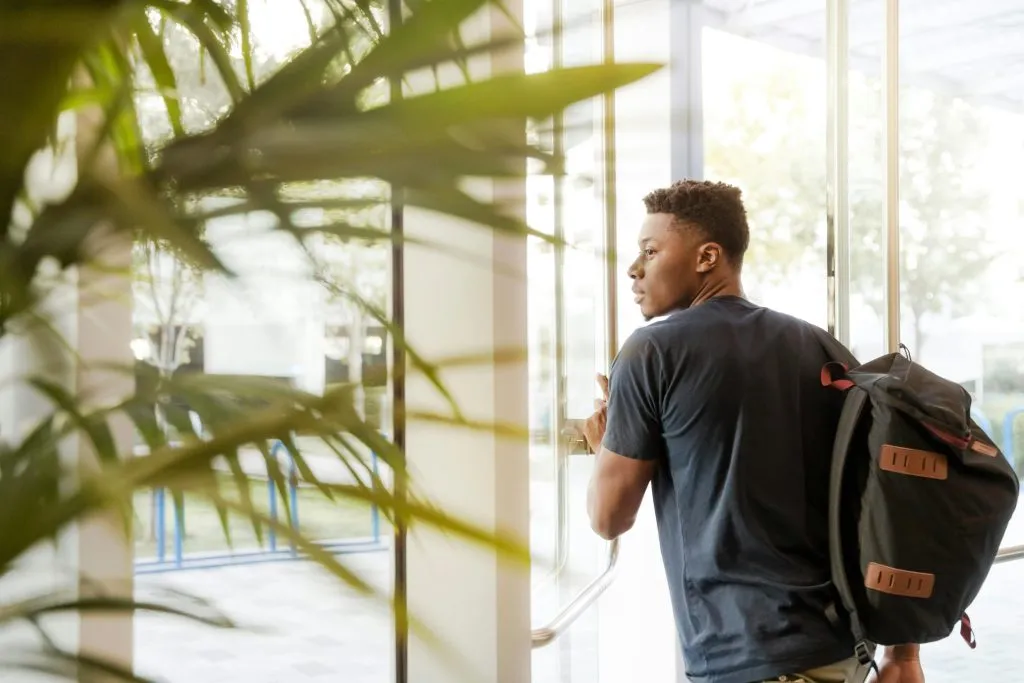 A man with a backpack, standing in a building with a plant.