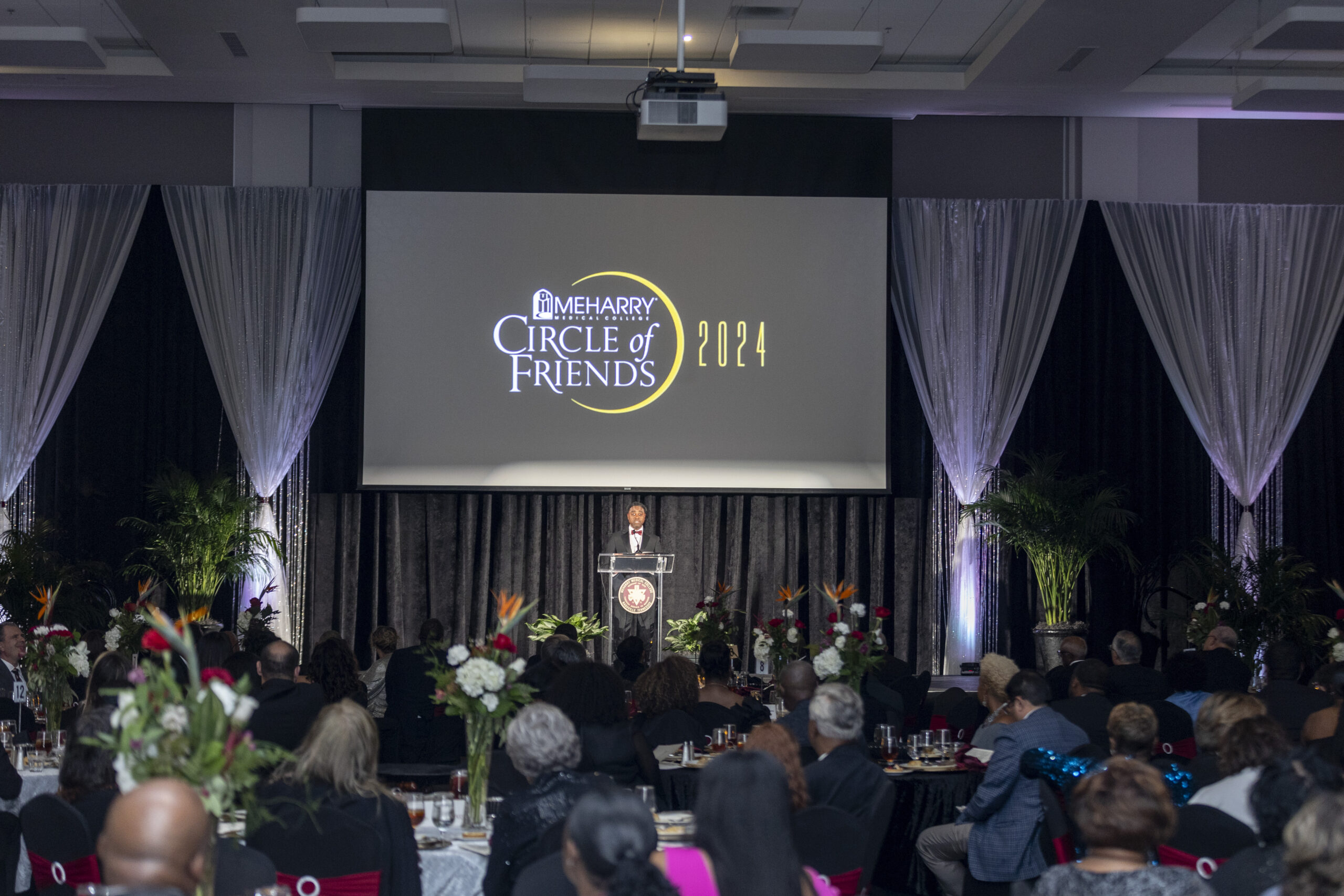 A large screen displaying a movie title, a speaker at a podium, and an audience seated in a dark room.