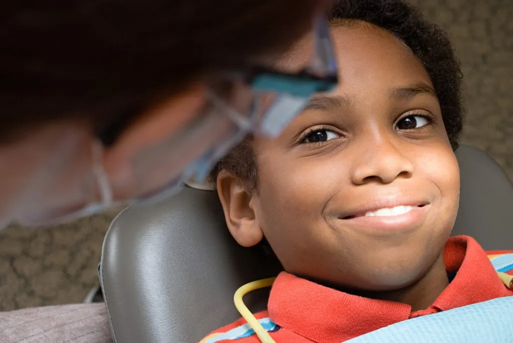 Young boy in dental chair, smiling, with adult's hands on his face.