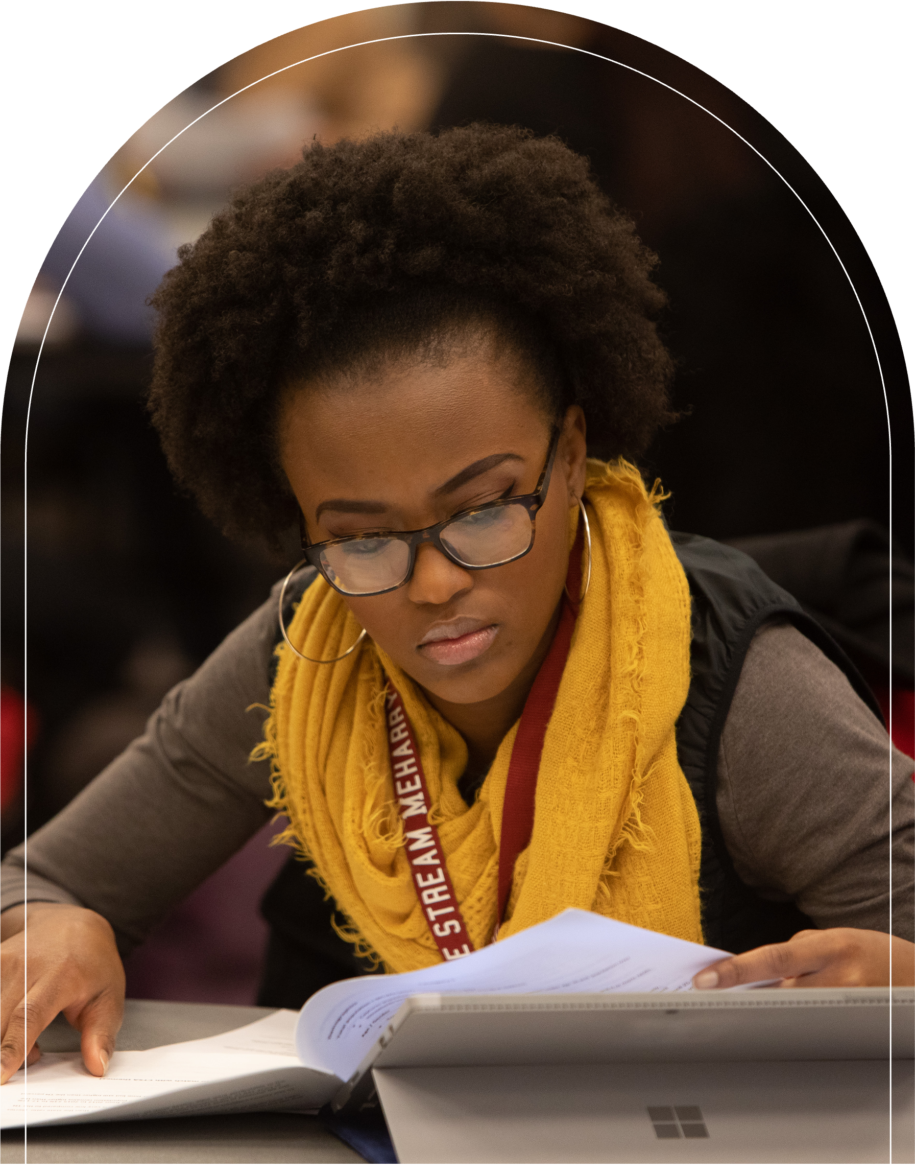 A woman in glasses, sitting at a table, reading a book. She has a laptop in front of her.