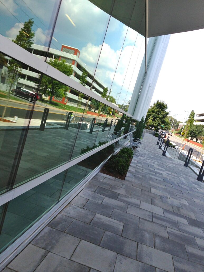 A modern building with a large glass wall, reflecting the outside. A sidewalk with a bench in front of the building.