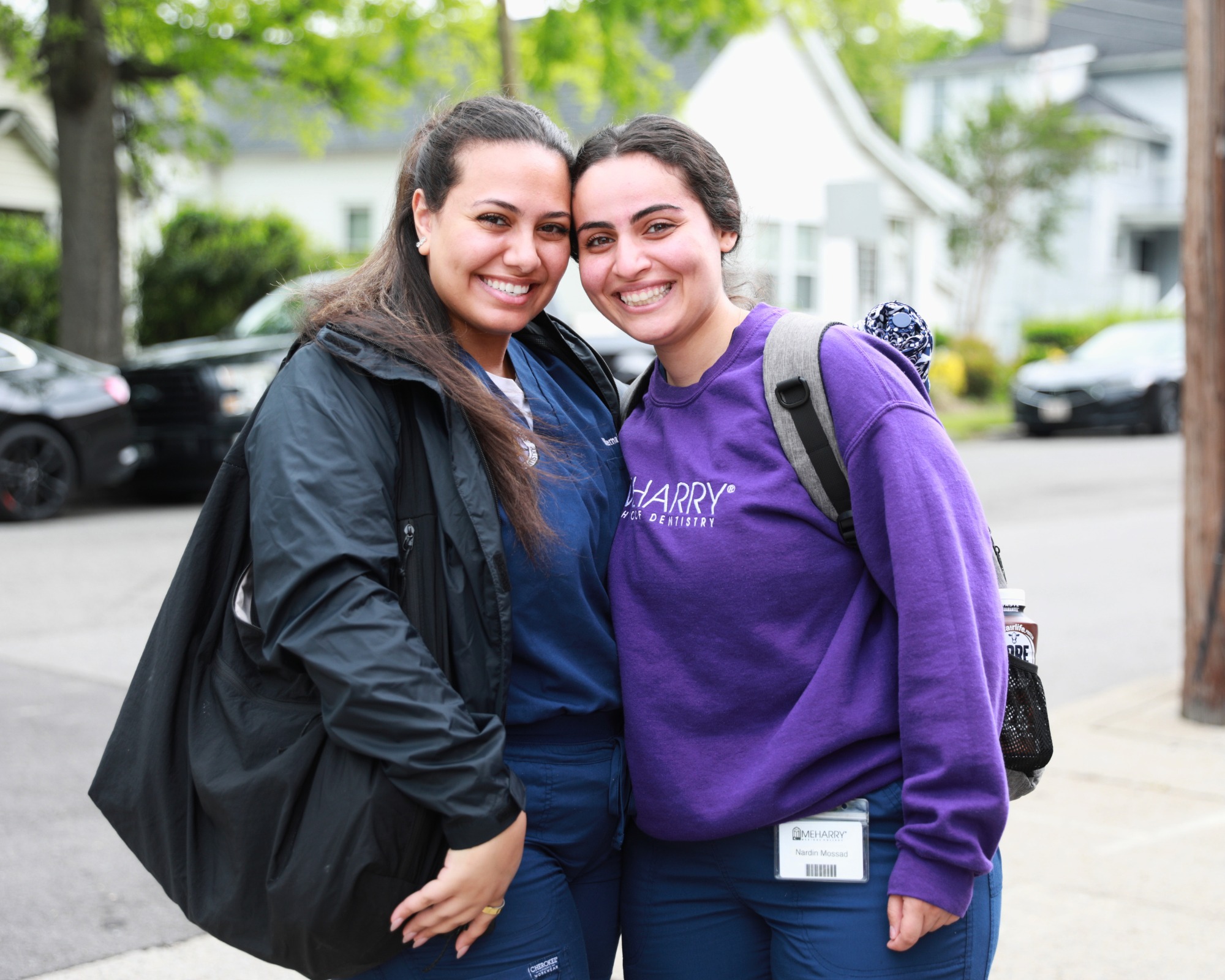 Two women posing for a photo, smiling, standing close together. One is wearing a blue jacket, the other a purple sweatshirt. They are in front of a house.