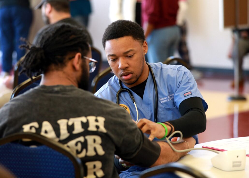 Two men in a room, one wearing a stethoscope, the other a nurse's cap. They are seated at a table with a laptop.