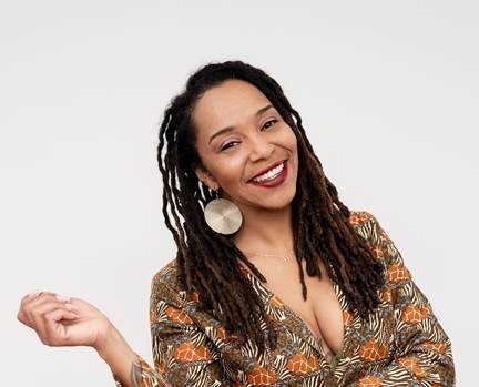 A woman with dreadlocks and a big smile, wearing a patterned top.