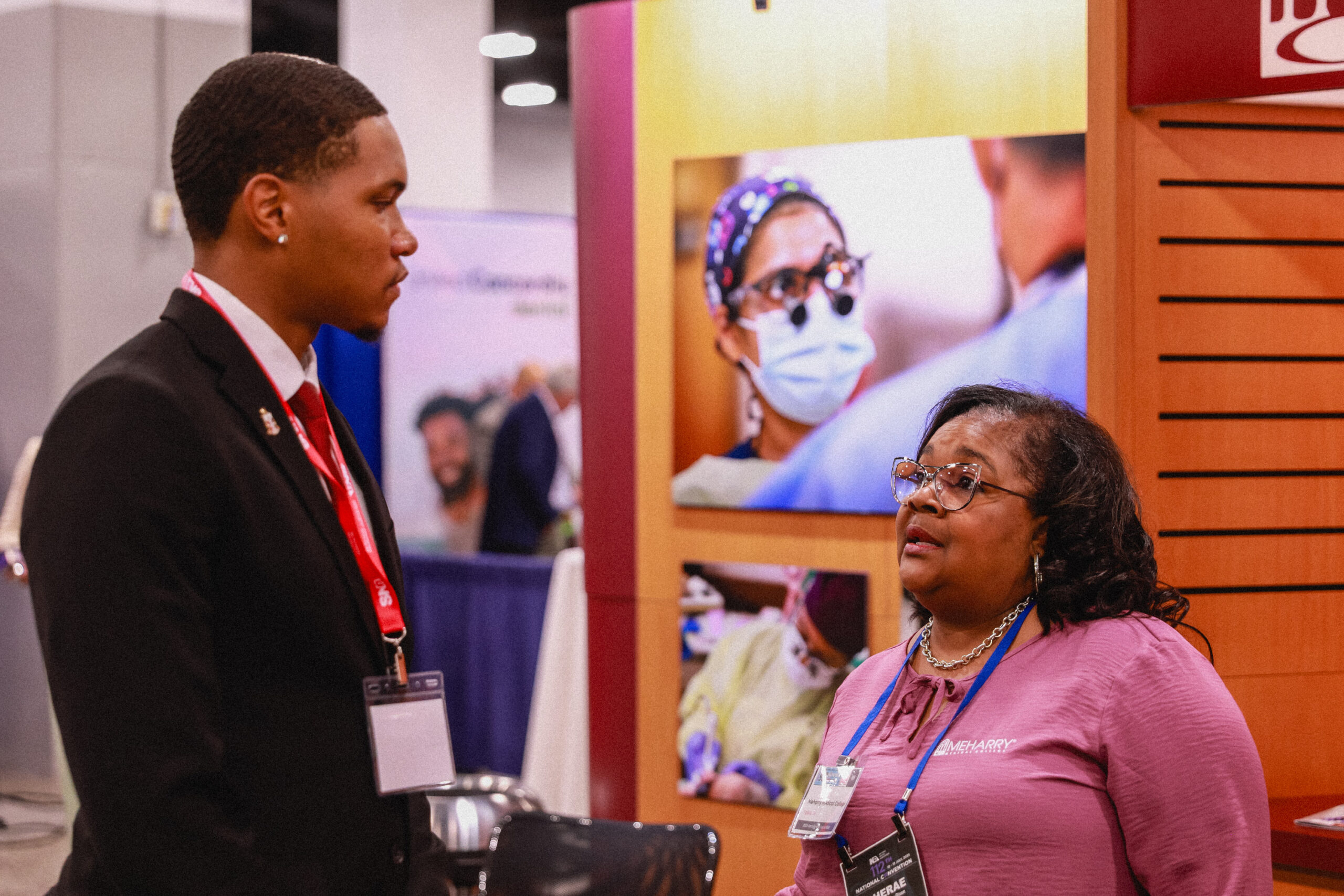 Two people at a booth, one man standing, one woman sitting, both wearing name tags.
