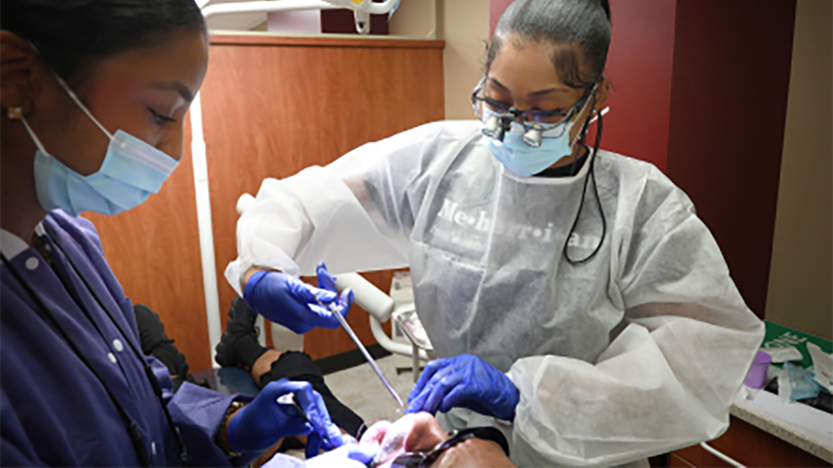 Two people in medical gowns, one wearing a mask, working on a patient.