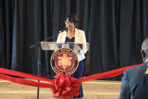 Woman speaking at podium, microphone, red ribbon, stage, audience.