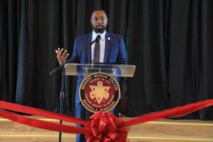 Man speaking at podium, microphone, red ribbon, black suit, white shirt, blue tie, name tag, audience, stage.