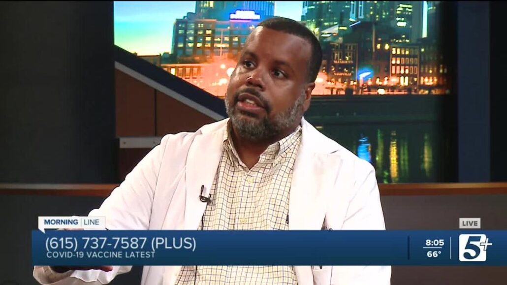Man sitting at news desk, holding microphone, smiling at camera.