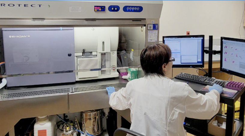 A person standing at a lab bench with a computer monitor and various lab equipment.