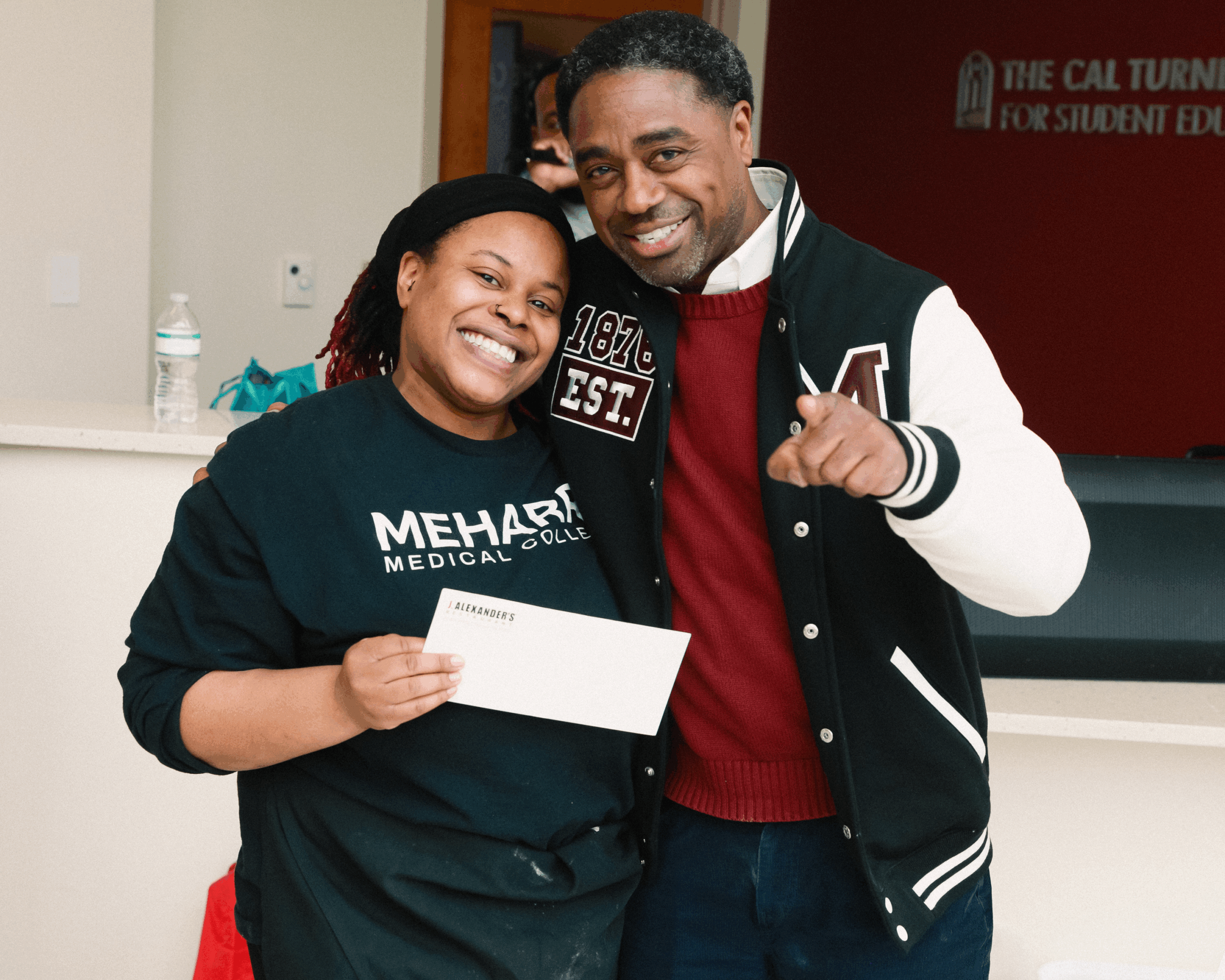 A man and woman posing for a photo. The man is holding a card. They are standing in front of a red wall with a white sign.
