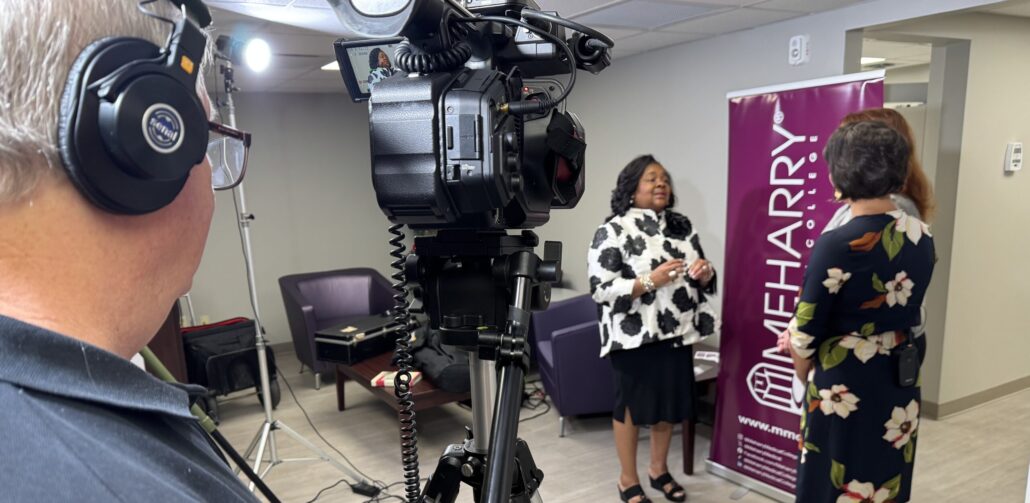A woman in a black and white dress standing in front of a camera. A man in a suit standing next to her. A professional video camera on a tripod. A news reporter recording the event. A woman in a floral dress standing in front of a sign. A man in a suit standing behind the woman. A news reporter recording the event.