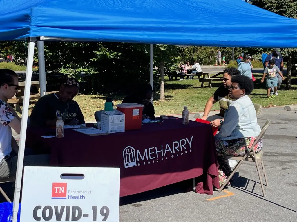 A blue tent with a table and chairs, people standing around, and a sign that reads "COVID-19 Vaccine Clinic".