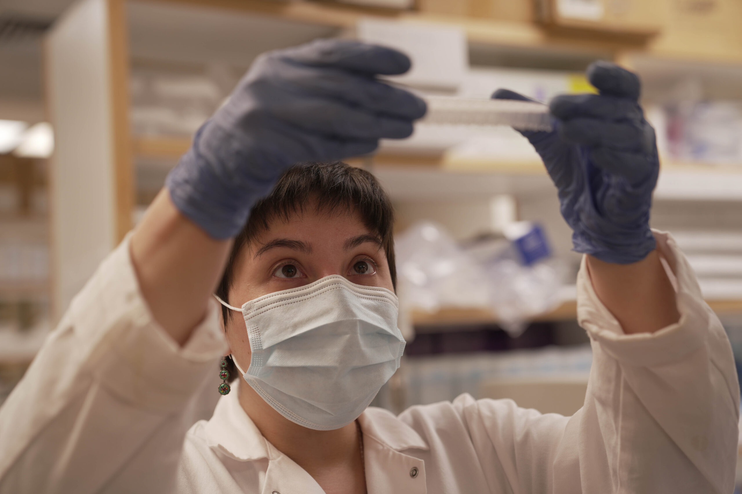Woman in lab coat and gloves holding a test tube.