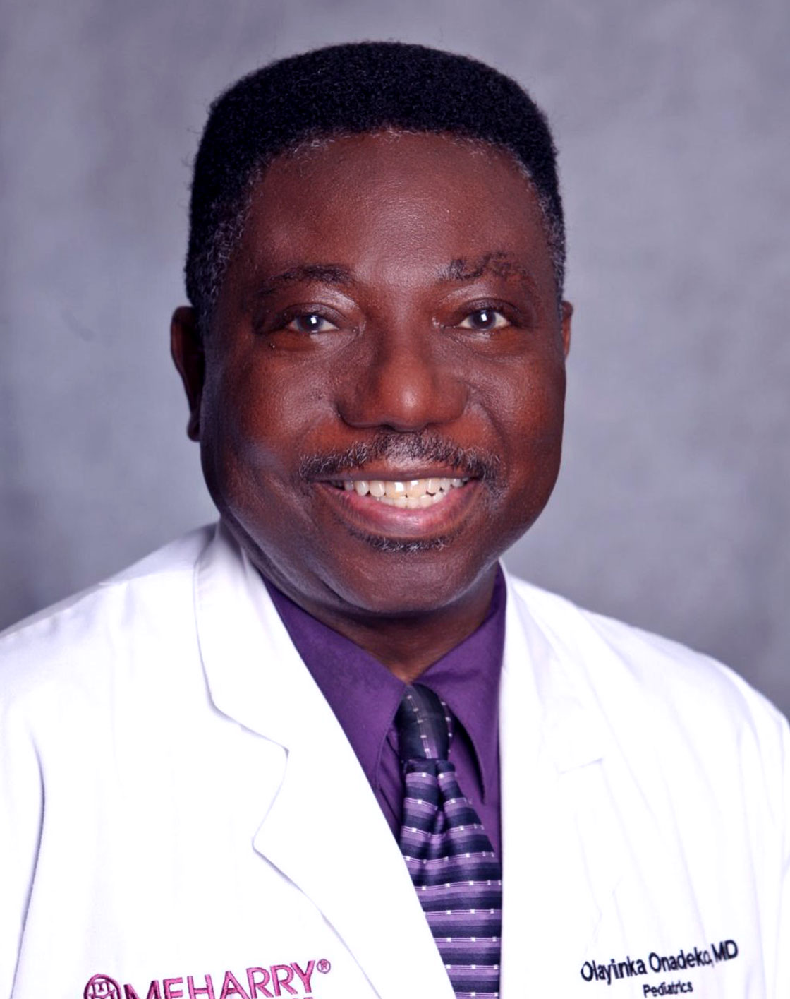 Man in white coat smiling, purple tie, purple shirt, balding, smiling.