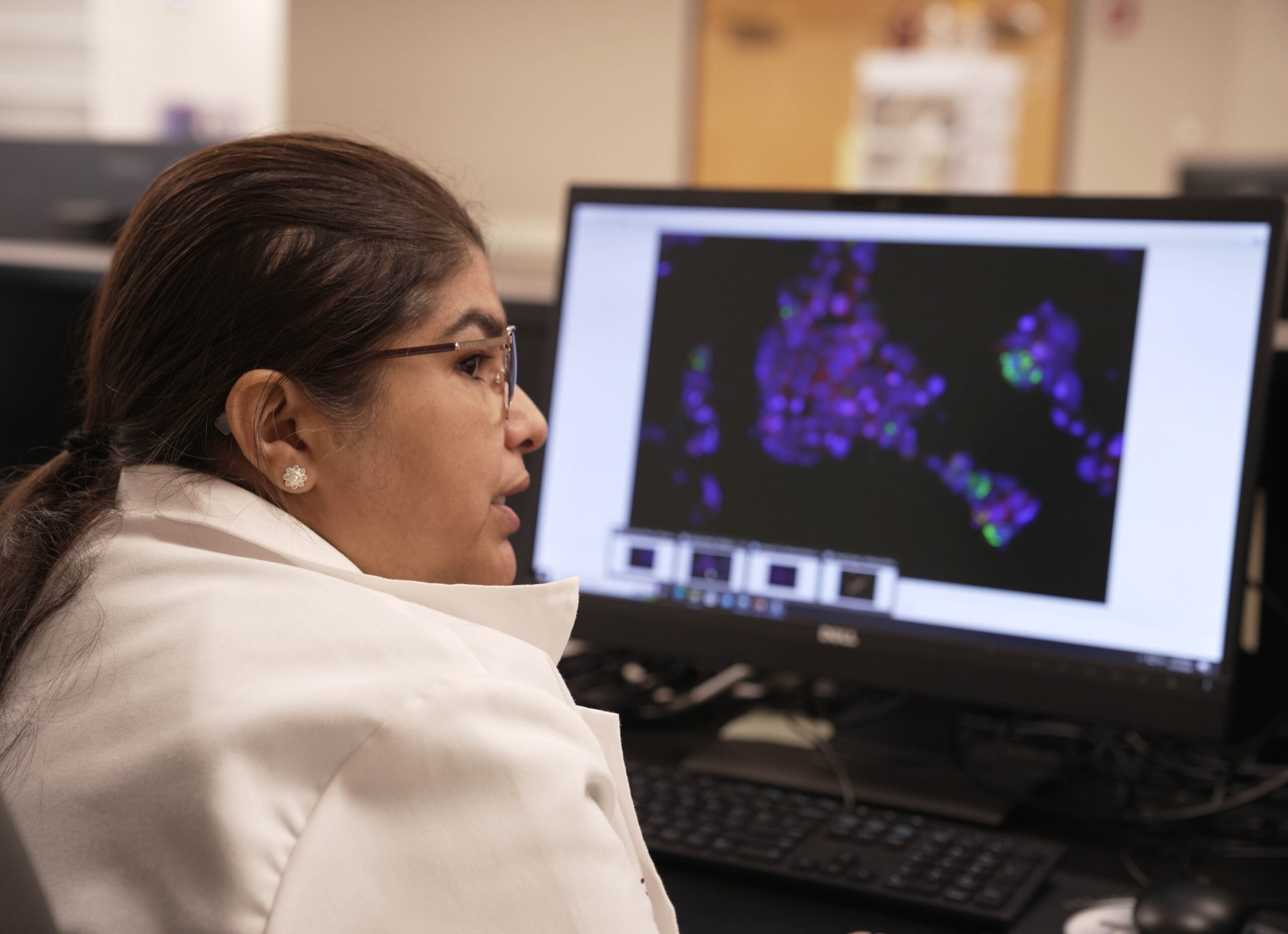 A woman sitting at a desk with a computer monitor displaying a colorful image. She is wearing glasses and a white coat.