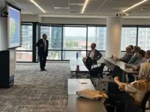 Man in suit standing in front of a presentation screen. People seated in a conference room, watching the presentation.