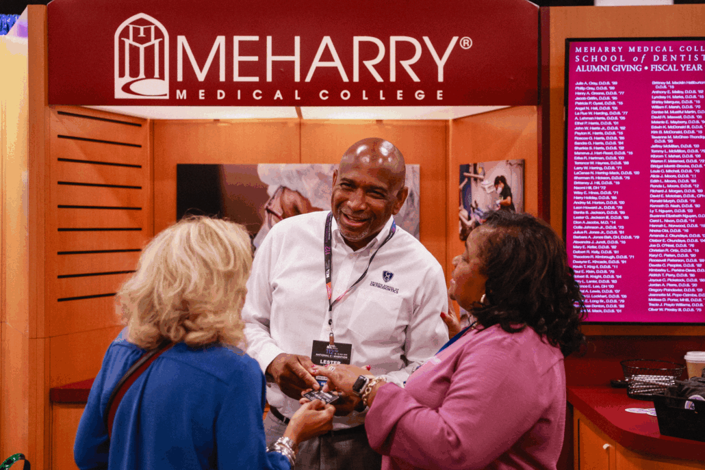Three people at a booth, one man standing, two women standing, all smiling. Sign behind them says "MEHARRY MEDICAL COLLEGE" and "MEHARRY MEDICAL COLLEGE".