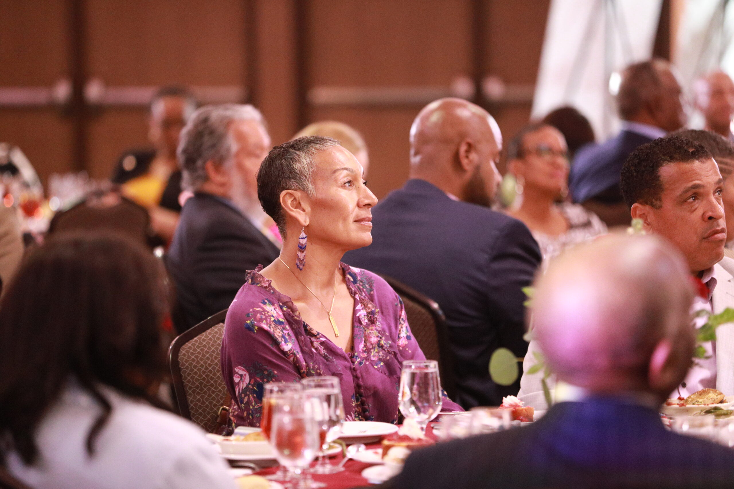 A group of people at a formal event, seated at a long table with food and drinks, engaged in conversation.