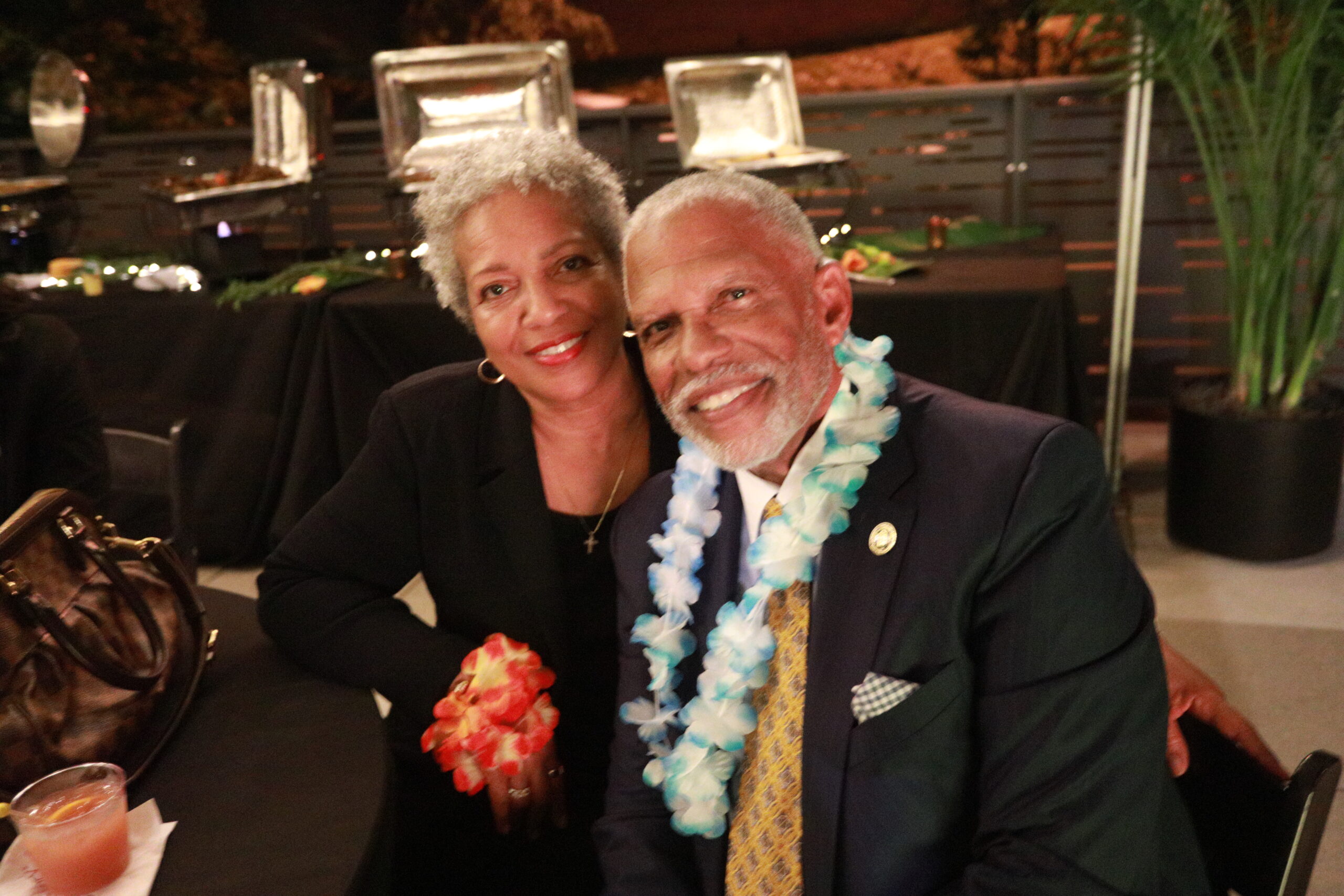 Couple posing for photo at event. Man in suit with lei around neck, woman in black.