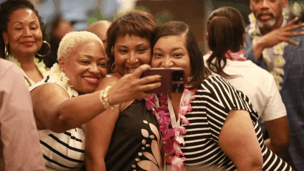 A group of people, including a woman in a black and white striped shirt, are taking a selfie together. They are at a social event, as indicated by the presence of leis.