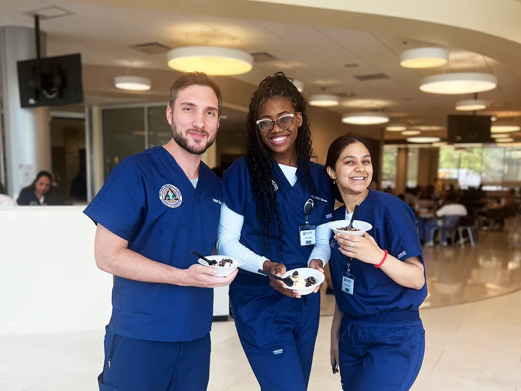 Three people in a room, posing with food. Two are holding plates, one is smiling. They are dressed in scrubs, suggesting they are nurses or doctors. The room has a modern, clean design, with a large window and a sign in the background.