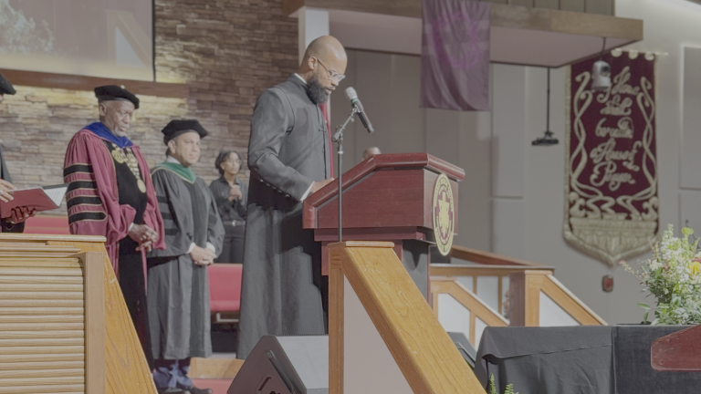 A man speaking at a podium, flanked by two men in robes.