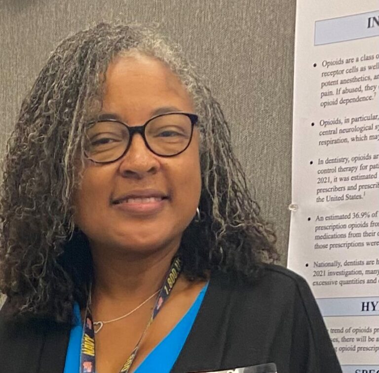 Woman with glasses, smiling, standing in front of a presentation board.
