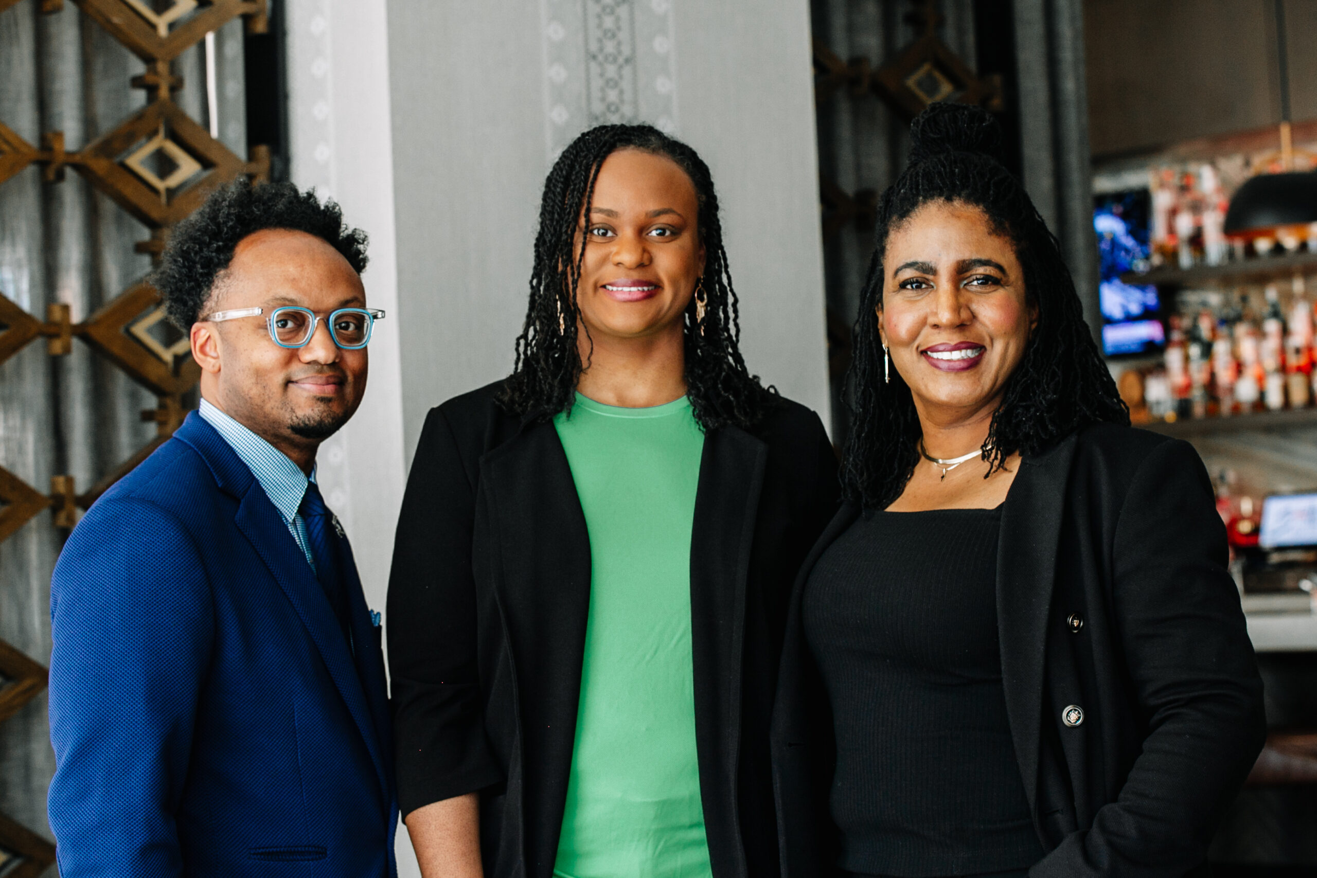 Three people posing for a photo. Woman in green shirt, man in blue suit, and woman in black shirt. They are standing in front of a bar.