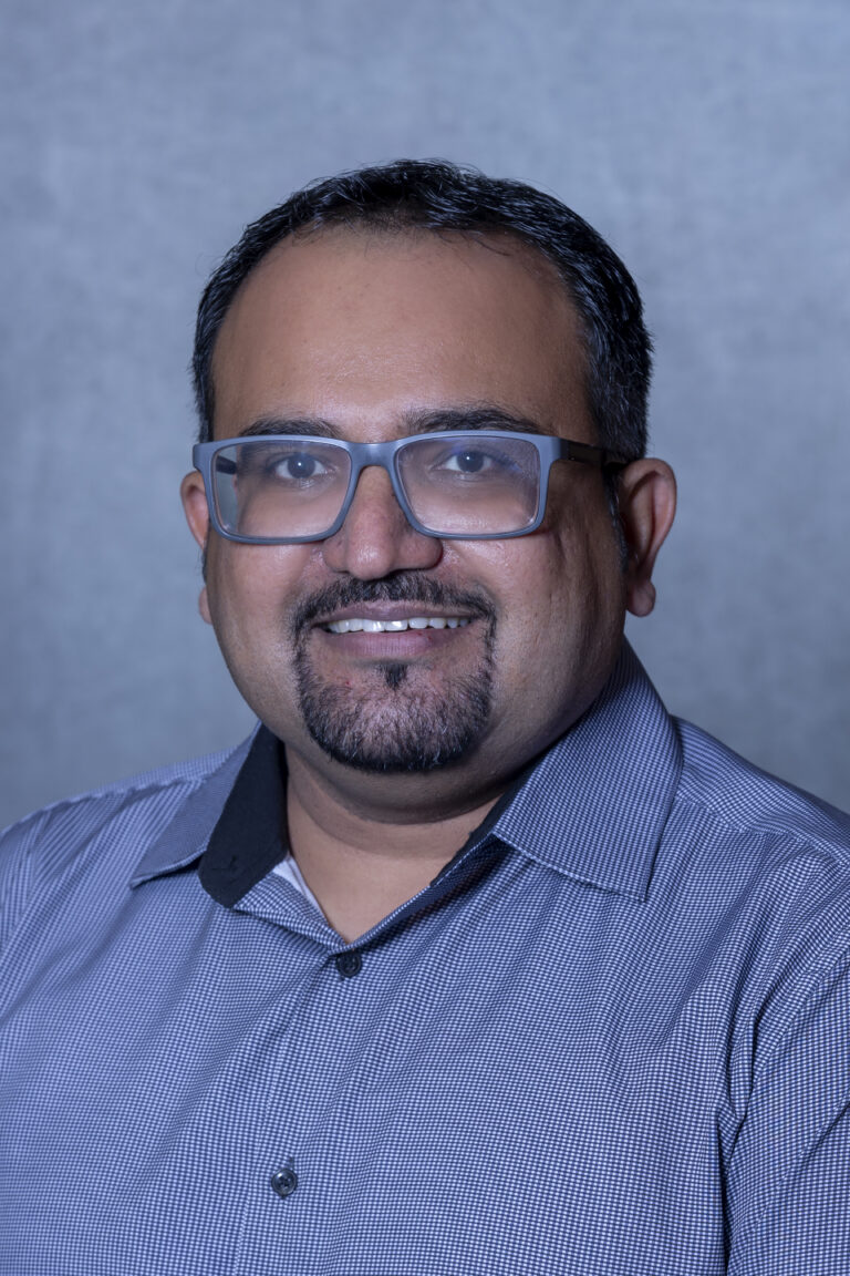 Man in glasses smiling, wearing glasses, suit jacket, and blue shirt.