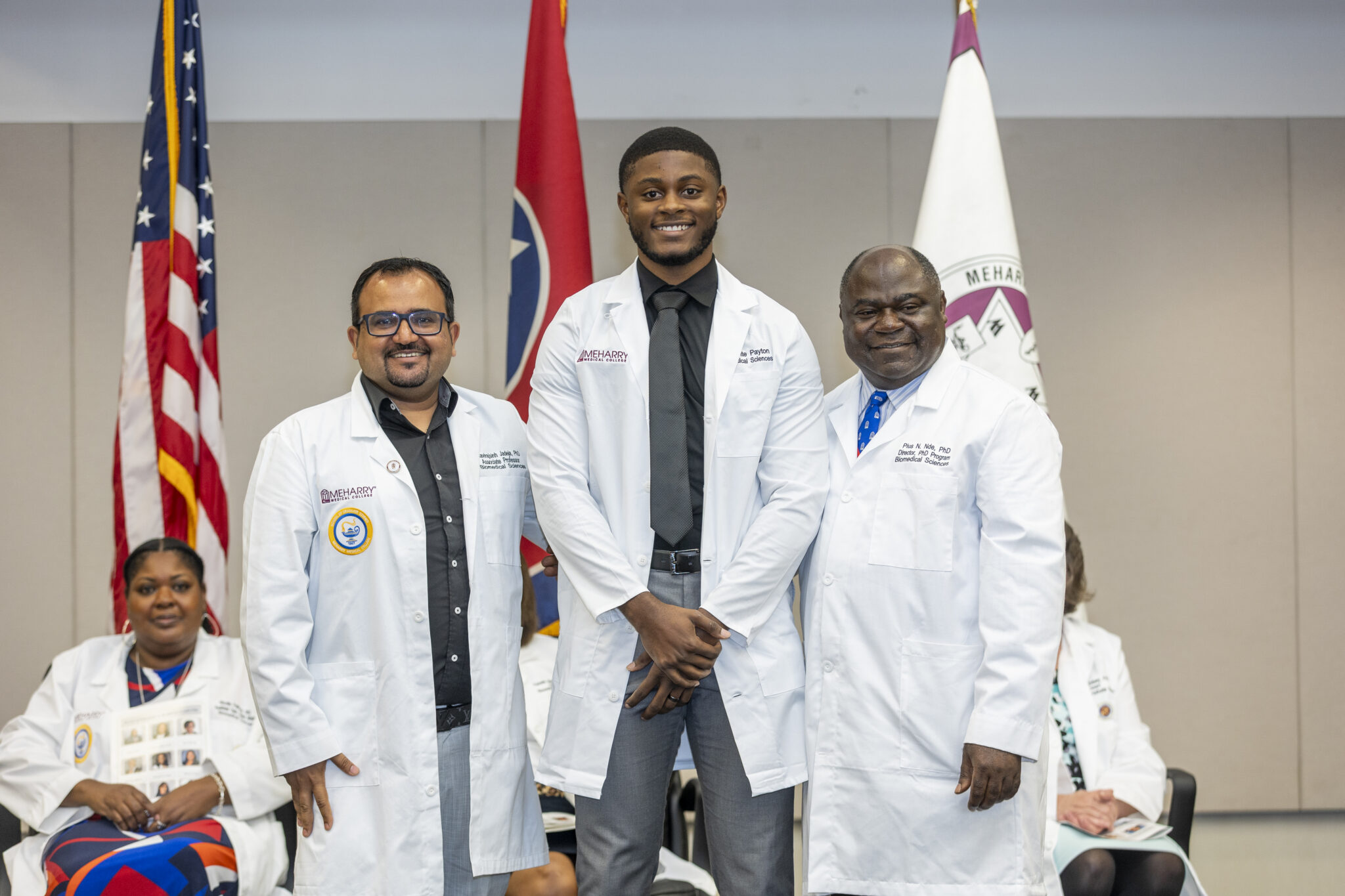 Three men standing together at a podium, one wearing a white lab coat. They are in front of flags and a person seated behind them.